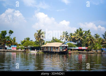 Alappuzha, Indien - 6. März 2023: Ein traditionelles Hausboot ist auf dem Nebenwasser in der Nähe mehrerer Gebäude am Fluss zu sehen, mit sichtbaren kommerziellen Schildern, darunter Coca-Cola-Werbung, mit Palmen und einem teilweise bewölkten Himmel im Hintergrund. Stockfoto