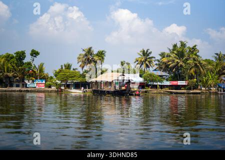 Alappuzha, Indien - 6. März 2023: Ein traditionelles Hausboot ist auf dem Nebenwasser in der Nähe mehrerer Gebäude am Fluss zu sehen, mit sichtbaren kommerziellen Schildern, darunter Coca-Cola-Werbung, mit Palmen und einem teilweise bewölkten Himmel im Hintergrund. Stockfoto