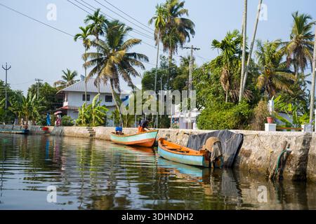 Alappuzha, Indien - 7. März 2023: Holzboote liegen entlang eines Kanals, der von Steinböcken umgeben ist, mit Palmen, Bananenpflanzen und Wohnhäusern sowie Menschen in der Nähe des Wassers unter klarem Himmel. Stockfoto