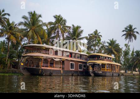 Alappuzha, Indien - 7. März 2023: Zwei große zweistöckige Hausboote aus Holz liegen nebeneinander am Wasser, umgeben von Palmen und tropischer Vegetation entlang der Ufer des Backwaters unter klarem Himmel. Stockfoto