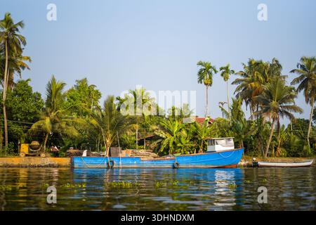 Alappuzha, Indien - 7. März 2023: Ein blaues Boot liegt entlang eines Kanals, der von Palmen, Bananenpflanzen und einem Steindamm mit Baumaterialien und Ausrüstungen umgeben ist, die am Ufer unter klarem Himmel sichtbar sind. Stockfoto