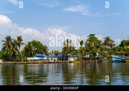 Alappuzha, Indien - 6. März 2023: Ein Gebäude am Flussufer mit sichtbarer „Devi Krishna“-Beschilderung und blauen Dächern ist entlang des Wassers zu sehen, mit Palmen und einem großen blauen Boot, das in der Nähe unter einem teilweise bewölkten Himmel angedockt ist. Stockfoto