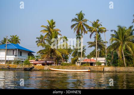 Alappuzha, Indien - 7. März 2023: Ein Kanal, der von einem Steindamm mit Palmen, Wohnhäusern und mehreren kleinen Booten umgeben ist, die am Wasser unter klarem Himmel vertäut sind. Stockfoto