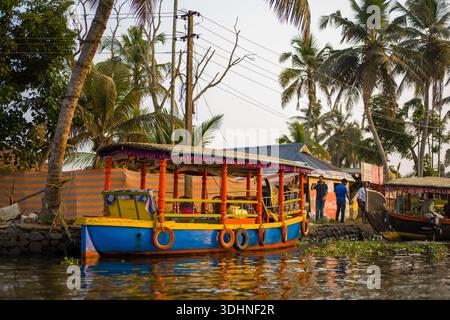 Alappuzha, Indien - 7. März 2023: Ein hell bemaltes, überdachtes Holzboot liegt entlang eines Kanals, auf dem mehrere Personen auf dem Steindamm in der Nähe von Palmen, Stromleitungen und Schildern unter klarem Himmel stehen. Stockfoto