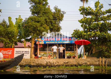 Alappuzha, Indien - 7. März 2023: Zwei Männer stehen vor dem Taste of India Backwater Restaurant, das ein prominentes Schild, Coca-Cola Branding und einen roten Regenschirm aufweist und von Bäumen am Ufer des Wassers umgeben ist. Stockfoto