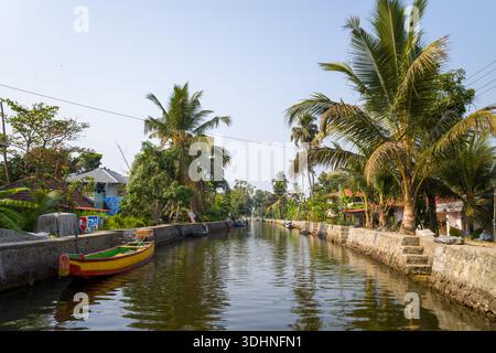 Alappuzha, Indien - 7. März 2023: Ein Kanal, gesäumt von Steinböcken, Palmen und Wohnhäusern, mit einem Holzboot, das am Wasserrand unter klarem Himmel vertäut ist. Stockfoto