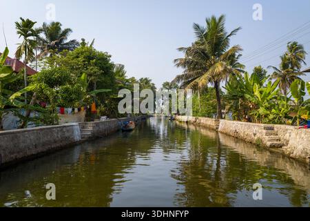 Alappuzha, Indien - 7. März 2023: Ein Kanal mit Steinböcken wird von Palmen, Wohnhäusern und vertäuten Booten begrenzt, wobei Wäsche hängt und Vegetation unter klarem Himmel sichtbar ist. Stockfoto