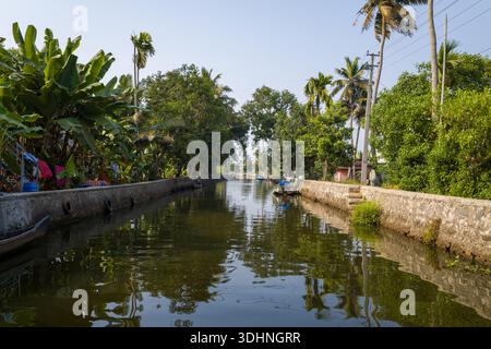 Alappuzha, Indien - 7. März 2023: Ein Kanal mit Steindämmen ist von Palmen und dichter Vegetation gesäumt, mit kleinen Booten, die entlang des Wassers vertäut sind, und Wäsche hängt auf der linken Seite unter klarem Himmel. Stockfoto