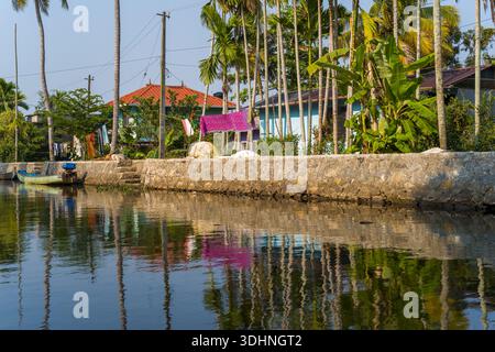 Alappuzha, Indien - 7. März 2023: Ein Kanal, der von einem Steindamm mit Wäscherei, Palmen, Bananenpflanzen und Wohnhäusern begrenzt wird, die unter klarem Himmel sichtbar sind. Stockfoto