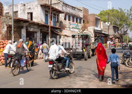Fatehpur Sikri, Indien - 12. März 2024: Das Bild zeigt eine lebhafte Straßenszene mit Menschen, die spazieren gehen, Fahrrad fahren und Motorräder fahren, und einen Traktor, der an alten Gebäuden und Marktständen in Fatehpur Sikri vorbeifährt. Stockfoto