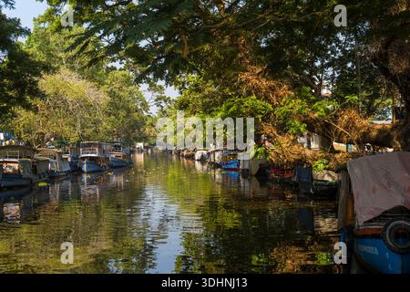 Alappuzha, Indien - 6. März 2023: Mehrere Boote liegen an beiden Seiten eines schmalen Kanals, der von großen Bäumen beschattet wird, mit Reflexionen auf dem Wasser und einer Brücke im Hintergrund. Stockfoto