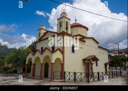 Stoupa, Griechenland - 10. September 2024: Das Bild zeigt das Äußere einer griechisch-orthodoxen Kirche mit cremefarbenen Wänden, roten Ziegeldächern, bogenförmigen Eingängen und Glockentürmen, die vor einem teilweise bewölkten Himmel stehen. Stockfoto