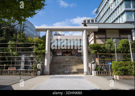 Tokio, Japan - 21. Oktober 2024: Das Bild zeigt den Vordereingang des Shiba Daijingu-Schreins mit einem großen Torii-Tor aus Stein, Steintreppen, die zum Hauptschrein führen, und moderne Bürogebäude im Hintergrund unter teilweise bewölktem Himmel. Stockfoto