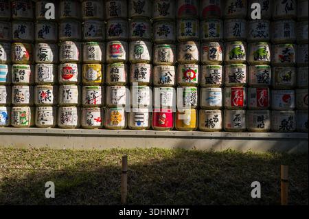 Tokio, Japan - 21. Oktober 2024: Reihen dekorativer Sake-Fässer mit farbenfroher japanischer Kalligraphie und Motiven werden im Freien am Meiji Jingu-Schrein ausgestellt, wobei das Sonnenlicht Schatten auf die Fässer wirft und Gras im Vordergrund steht. Stockfoto