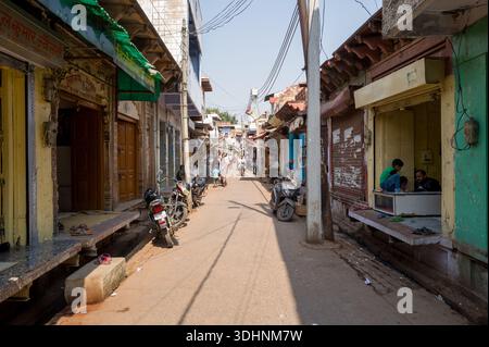 Fatehpur Sikri, Indien - 12. März 2024: Das Bild zeigt eine enge Straße mit kleinen Geschäften, seitlich geparkten Motorrädern, Freileitungen und mehreren Personen, die unter Tageslicht sitzen oder gehen. Stockfoto