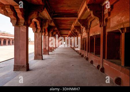 Fatehpur Sikri, Indien - 12. März 2024: Das Bild zeigt einen langen roten Sandsteinkorridor mit Säulenreihen und geschnitzten Klammern, Teil des Stalls im Jodha Bai Palace in Fatehpur Sikri, mit offenen Ständen und einem leeren Innenhof. Stockfoto