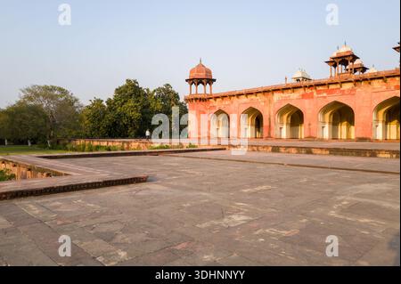 Agra, Indien - 12. März 2024: Das Bild zeigt eine Seitenansicht der roten SandsteinArkade mit bogenförmigen Eingängen und einem erhöhten Pavillon mit Chhatri im Akbar's Tomb Complex, mit einer Person, die in der Nähe der Bäume spaziert, und einem offenen gepflasterten Innenhof im Vordergrund unter klarem Himmel. Stockfoto
