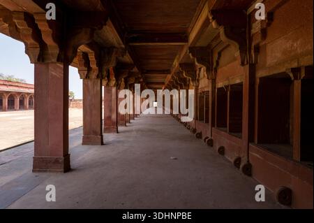 Fatehpur Sikri, Indien - 12. März 2024: Das Bild zeigt einen langen roten Sandsteinkorridor mit Säulenreihen und geschnitzten Klammern, Teil des Stalls im Jodha Bai Palace in Fatehpur Sikri, mit offenen Ständen und einem leeren Innenhof. Stockfoto