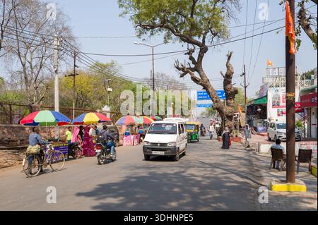 Fatehpur Sikri, Indien - 12. März 2024: Das Bild zeigt eine belebte Straße mit bunten Marktständen, Fußgängern, Fahrrädern, Motorrädern, Autos, und Auto-Rikschas in der Nähe des Eingangs zu Fatehpur Sikri unter klarem Himmel. Stockfoto