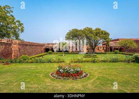 Fatehpur Sikri, Indien - 12. März 2024: Das Bild zeigt einen Landschaftsgarten mit Blumenbeeten, getrimmten Rasenflächen und Reifen Bäumen, umgeben von roten Sandsteinmauern und bogenförmigen Kolonnaden im Jodha Bai Palace Komplex in Fatehpur Sikri. Stockfoto