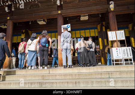 Tokio, Japan - 21. Oktober 2024: Menschen, darunter ein uniformierter Sicherheitsbeamter, werden auf den Stufen des Haupteingangs des Meiji Jingu-Schreins stehend und gehen gesehen, mit Spendenboxen, Informationsschildern und traditioneller Holzarchitektur. Stockfoto