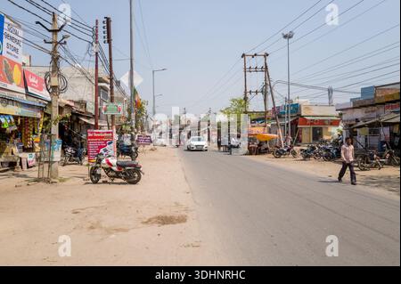 Mathura, Indien - 13. März 2024: Das Bild zeigt einen weiten Blick auf eine Hauptstraße in Mathura mit verschiedenen Geschäften, Motorrädern am Straßenrand, Fußgängern, einem weißen Auto, und Freileitungen bei klarem Tageslicht. Stockfoto