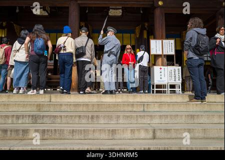 Tokio, Japan - 21. Oktober 2024: Eine Gruppe von Menschen, darunter ein uniformierter Sicherheitsbeamter, wird an den Eingangspunkten des Meiji Jingu-Schreins stehend und gehen gesehen, mit Spendenboxen und Informationsschildern, die vor dem hölzernen Schrein sichtbar sind. Stockfoto