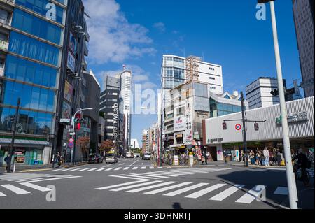 Tokio, Japan - 21. Oktober 2024: Das Bild zeigt eine geschäftige Kreuzung in Shibuya mit Fußgängern, die an Straßenübergängen warten, modernen Glasgebäuden, älteren Flachbauten, sichtbaren Schildern und einem klaren blauen Himmel. Stockfoto