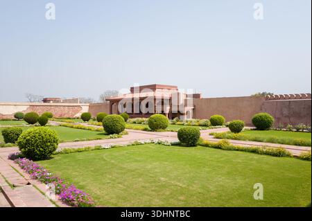 Fatehpur Sikri, Indien - 12. März 2024: Das Bild zeigt den roten Sandstein Diwan-E-AAM-Publikumssaal des Jodha Bai Palastkomplexes mit gepflegten Rasenflächen, Blumenbeeten und getrimmten Büschen im Vordergrund unter klarem Himmel. Stockfoto