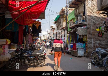 Fatehpur Sikri, Indien - 12. März 2024: Ein Mann läuft eine enge Straße entlang, die von kleinen Geschäften gesäumt ist, Motorräder an den Seiten parken, hängende Textilien, Drähte und mehrere Personen, die im Hintergrund bei Tageslicht sichtbar sind. Stockfoto