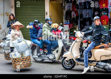 Ein alter vietnamesischer Händler trägt Bambushut, trägt panier mit Utensilien, spaziert an vierköpfiger Familie vorbei, beim Motorradeinkaufen in der Altstadt Stockfoto