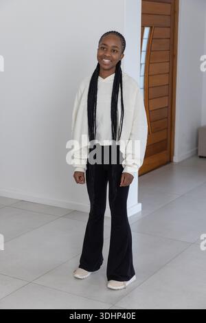 Afroamerikanische Frau, die im Eingang durch eine Holztür auf gefliestem Boden steht und einen cremefarbenen Hoodie trägt Stockfoto