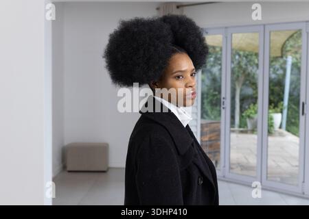 Afroamerikanerin, die in einem hellen Raum neben Schiebetüren steht, in schwarzem Mantel und weißem Hemd Stockfoto