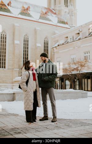 Eine Koreanerin mit beigefarbenem Mantel und rotem Schal steht an einem schneebedeckten Tag mit einem kaukasischen Mann in grünem Mantel. Sie stehen vor einer Kirche in der EU Stockfoto