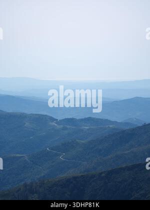 Überlagerte Bergkämme auf Sardinien verblassen sich unter einem trüben Himmel in die Ferne, während sich eine Serpentinenstraße durch die Hügel schlängelt. Weiches Licht. Stockfoto