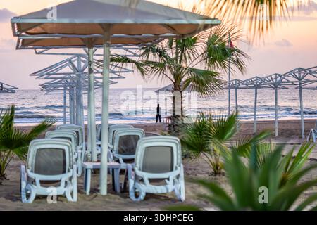 Ruhige Strandszene mit weißen Liegestühlen und Sonnenschirmen bei Sonnenuntergang, mit einer einsamen Silhouette am Meer in Side, Antalya. Stockfoto