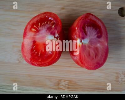 Top-Ansicht der frischen Reifen Tomaten, die auf einem Holzschneidebrett in zwei Hälften geschnitten wurden, mit saftiger Textur, Rezepten und Food-Blog-Inhalten für Küchen- und Kochkünstler Stockfoto