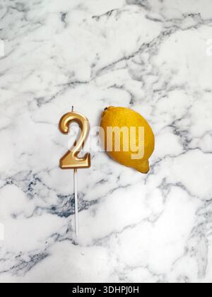 Auf der Marmoroberfläche liegen eine goldene Kerze in Form der Zahl 2 und eine Zitrone. Komposition, 2 Millionen, Blogger, Feier, Abonnenten Stockfoto