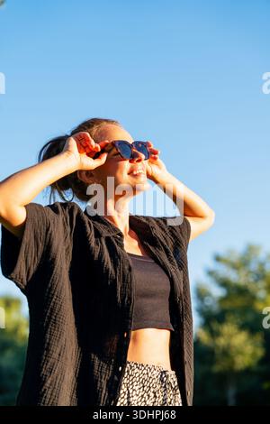 Lässiges Porträt einer jungen erwachsenen Frau mit schwarzem Outfit und Sonnenbrille bei warmem Sonnenlicht. Stockfoto