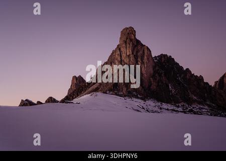 Wintersonnenuntergang am Passo Giau in den Dolomiten, wo warme alpine Lichter mit schneebedeckten Gipfeln unter einem dramatischen violetten Himmel kontrastieren. Stockfoto