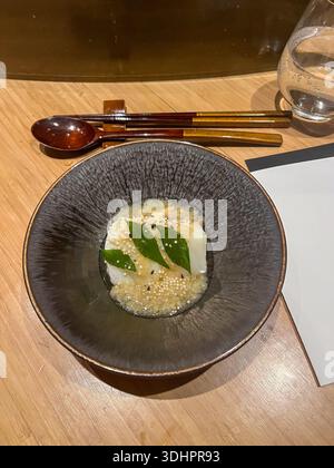 Ein zarter weißer Block, möglicherweise Tofu oder Panna Cotta, ruht in einer schimmernden Sauce, garniert mit grünen Blättern und winzigen Samen, in einer dunklen Schüssel. Stockfoto