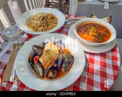 Drei appetitliche Gerichte, darunter Nudeln mit Meeresfrüchten, gedämpfte Muscheln und eine herzhafte Suppe, werden auf einem Tisch im Freien serviert. Stockfoto