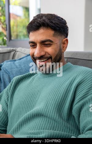 Erwachsener indischer Mann sitzt auf grauem Sofa und lächelt in blauem Pullover mit blauem Kissen, Blick auf das Fenster Stockfoto