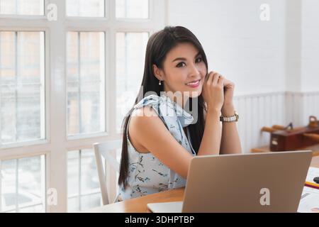 Portrait einer lächelnden, hübschen Geschäftsfrau, die auf einem Holzschreibtisch im Büro sitzt und die Kamera betrachtet. Stockfoto