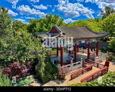 Ein kunstvoller traditioneller ostasiatischer Pavillon mit roten Säulen und farbenfrohen Details steht in einem üppig grünen Garten unter einem sonnigen blauen Himmel. Stockfoto