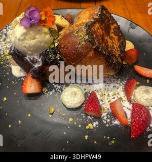 Ein elegantes Dessert besteht aus goldenen French Toast Scheiben, schmelzendem Vanilleeis und frischem Obst auf einer dunklen Keramikplatte. Stockfoto