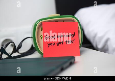 Ein roter Haftnotiz mit dem Aufdruck Guten Morgen, den man über einem grünen Wecker auf einem Nachttisch mit einer Brille und einem geschlossenen Tablet im Vordergrund hatte Stockfoto