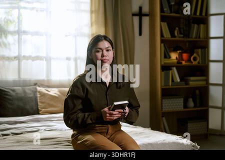 Porträt einer jungen asiatischen Frau, die drinnen sitzt und die Bibel hält Stockfoto