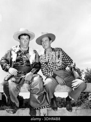 Aus den 1940er Jahren zeigt dieses Schwarz-weiß-Duo noch immer das legendäre Comedy-Duo Abbott und Costello, das in Cowboykleidung gekleidet ist und mit Strohhalmen im Mund auf einem Zaun sitzt. Lou Costello (links) trägt ein Satinhemd mit einer Pelzweste und Chaps, während Bud Abbott (rechts) ein kariertes Hemd und einen Schal trägt, sowohl nach oben als auch nach rechts, auf einem Filmset für ihren Film „Ride 'EM Cowboy“. Stockfoto