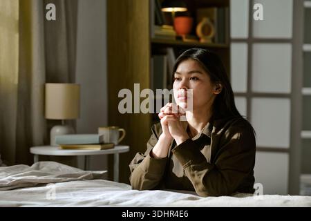Junge Erwachsene asiatische Frau, die auf dem Bett sitzt und sich in einem hellen, modernen Schlafzimmer spiegelt Stockfoto